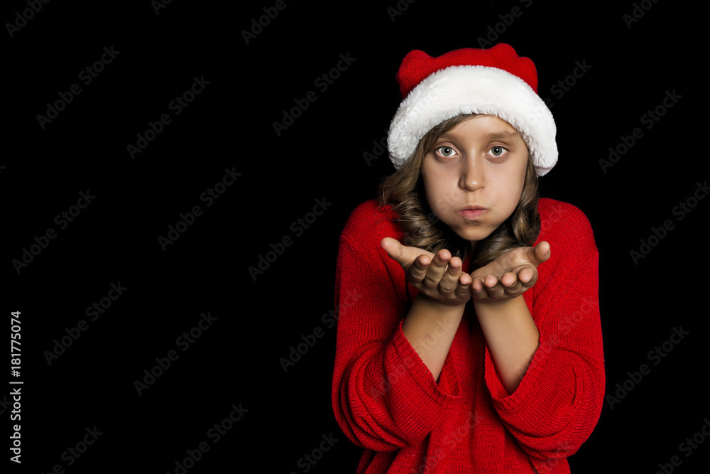 A young girl in a red sweater and a Santa Claus hat blows on her hands and looks at the viewer A young girl in a red sweater and a Santa Claus hat blows on her hands and looks at the viewer