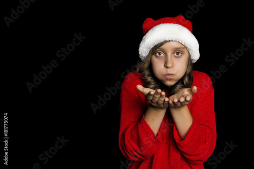 A young girl in a red sweater and a Santa Claus hat blows on her hands and looks at the viewer