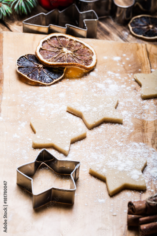Christmas gingerbread cookies in the making on wooden cutting board, pastry dough star shape cutters. gift boxes red ribbon, rolling pin, cinnamon sticks orange slices red potholder Christmas gingerbread cookies in the making on wooden cutting board, pastry dough star shape cutters. gift boxes red ribbon, rolling pin, cinnamon sticks orange slices red potholder