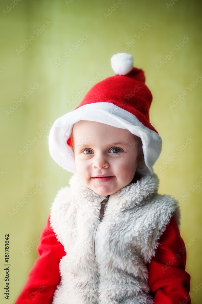 A baby dressed as Santa Claus with a red cap on his head and a white fur vest looks and smiles. A baby dressed as Santa Claus with a red cap on his head and a white fur vest looks and smiles.