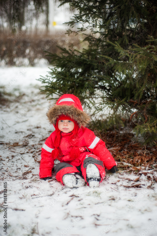 A baby in a red jumpsuit sits on the snow under a green tree and looks