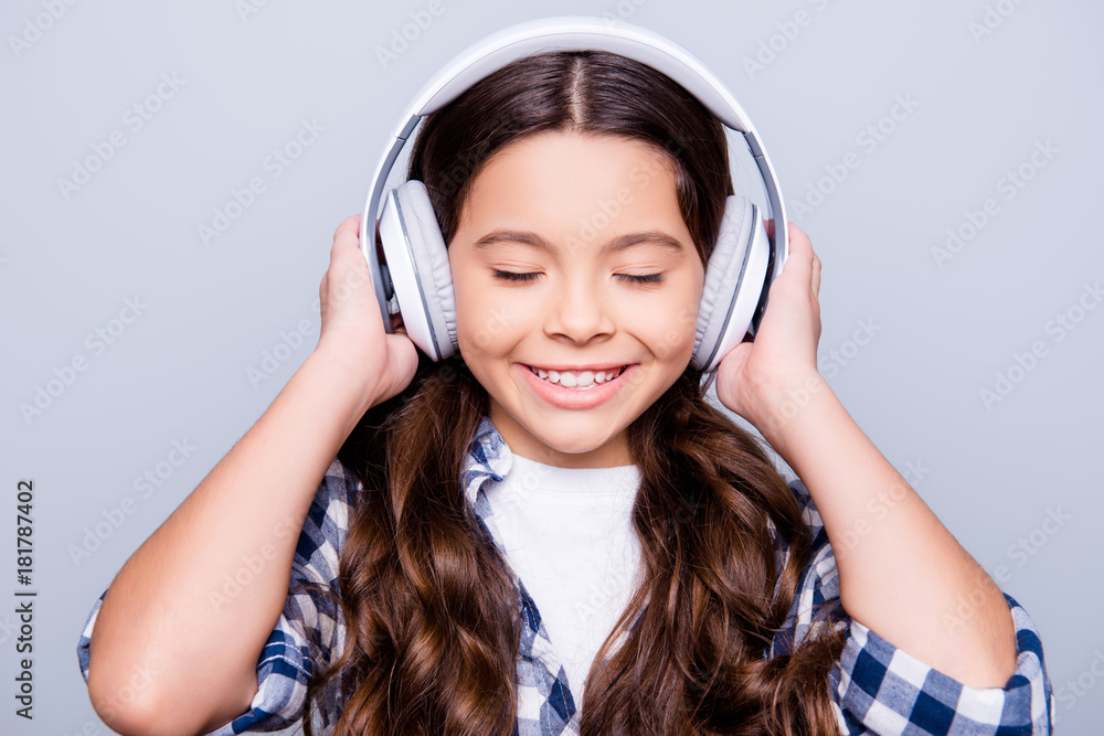 Close up portrait of brunette charming smiling kid holding headphones on her head enjoying her favorite music with close eyes standing over grey background Close up portrait of brunette charming smiling kid holding headphones on her head enjoying her favorite music with close eyes standing over grey background