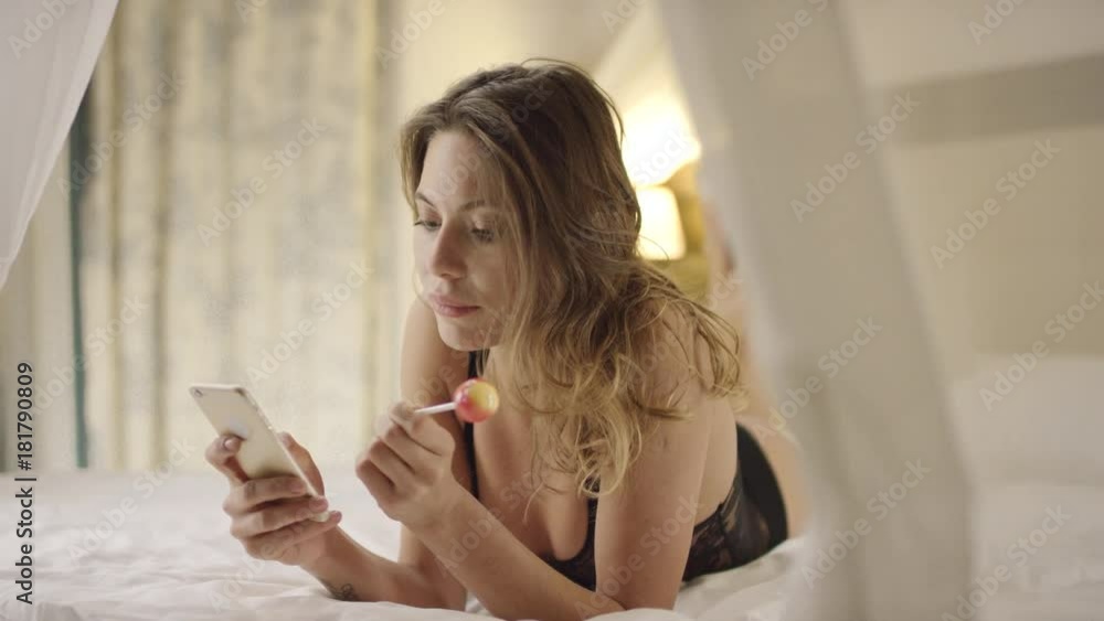 Charming woman using smartphone and licking hard candy on bed Charming woman using smartphone and licking hard candy on bed