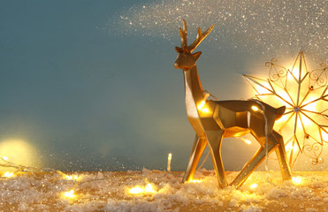 Gold shiny reindeer on snowy wooden table with christmas garland lights.