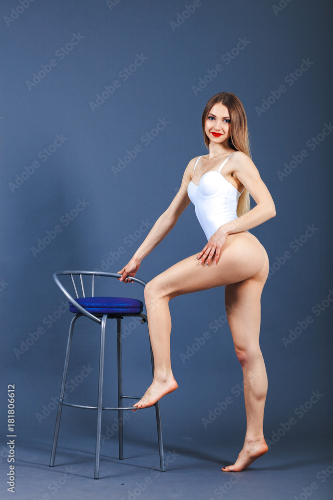 Young happy fitness girl with sporty body posing at studio on a blue background. Beautiful fit Girl. Fitness model in white sports bodysuit stands next to chair. Healthy lifestyle. Sexy woman.