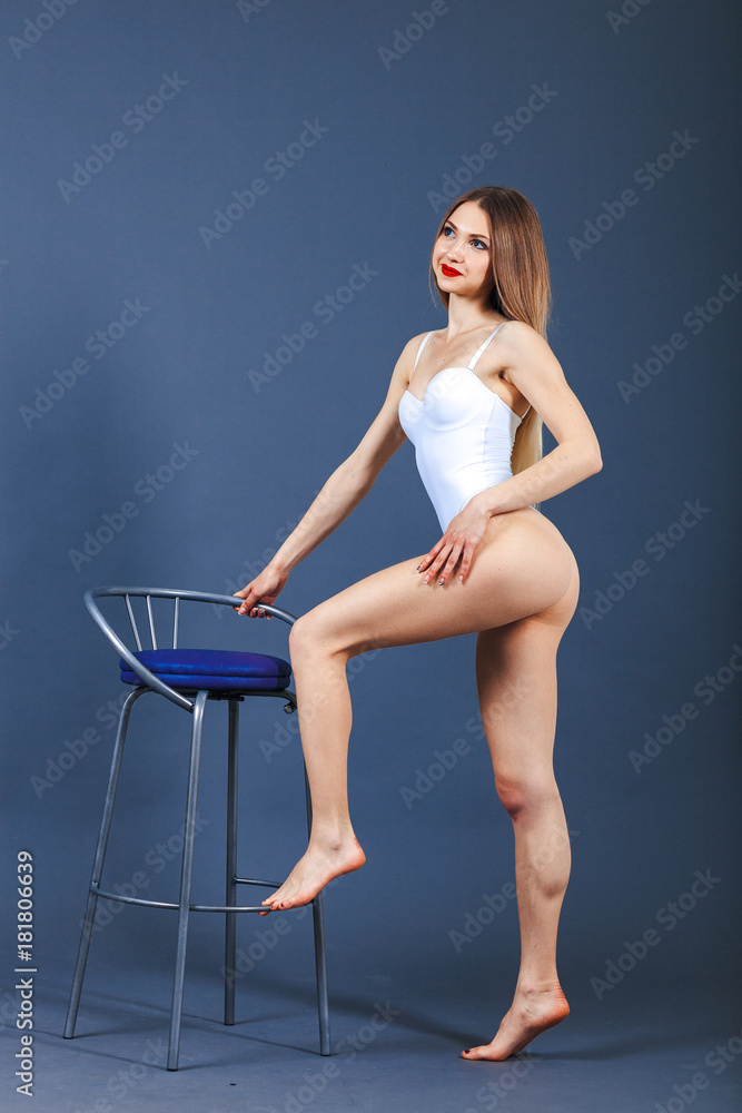 Young happy fitness girl with sporty body posing at studio on a blue background. Beautiful fit Girl. Fitness model in white sports bodysuit stands next to chair. Healthy lifestyle. Sexy woman.
