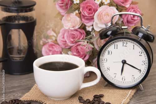 Hot cup of coffee and alarm clock on wood table with rose flower.