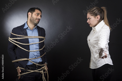 un uomo colpevole in giacca legato con una corda viene guardato da una moglie furiosa - sfondo scuro 