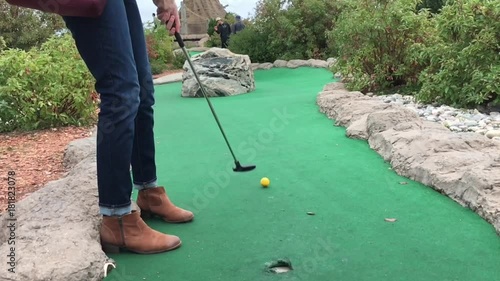 Lower part of woman wearing boots hits yellow golf ball in slow motion on mini golf course.  
