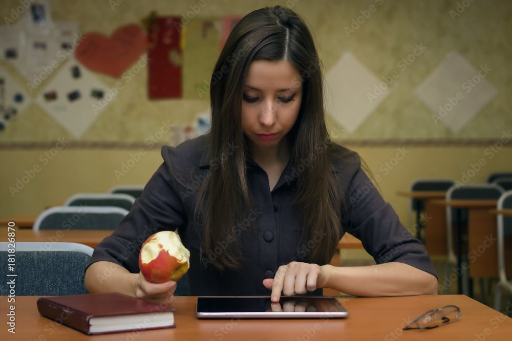 Caucasian young female student girl is sitting at a desk in the school classroom holds and eats an apple and reads lecture in her tablet computer pc or looking for information. Education concept. Caucasian young female student girl is sitting at a desk in the school classroom holds and eats an apple and reads lecture in her tablet computer pc or looking for information. Education concept.
