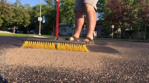 Man sweeping with push broom in angled sunlight moves sand on dark surface towards ground based camera.  