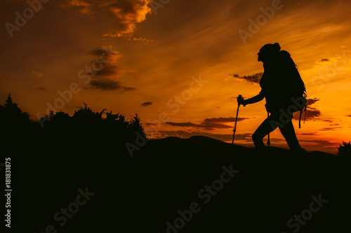 Silhouette hiker woman tracking with backpack and trekking pole, sunset orange sky on the background