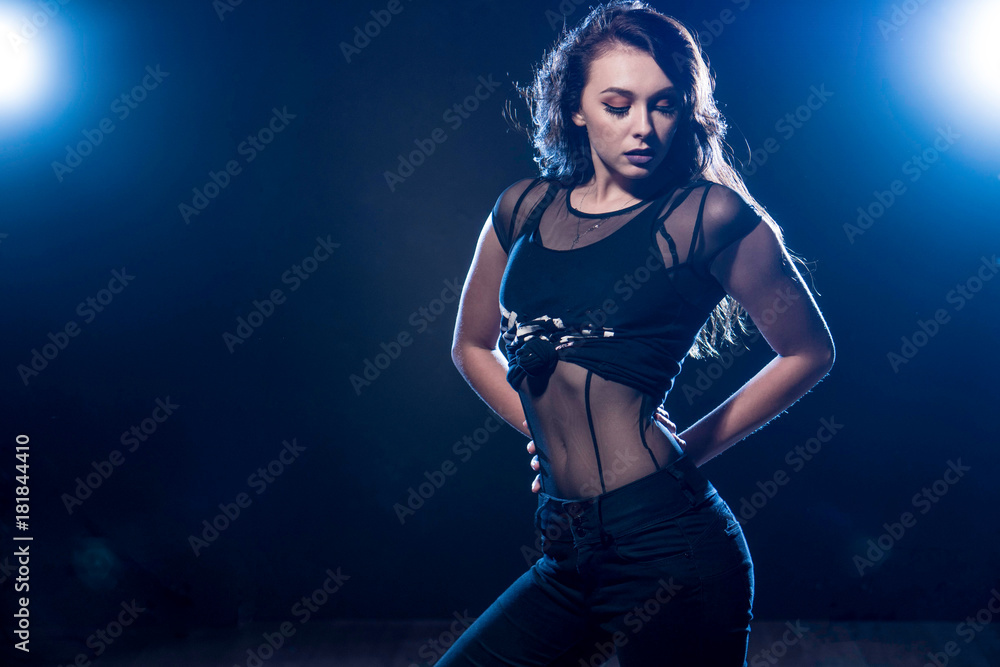 young adult woman standing in lights wearing black bodysuit, and jeans.
