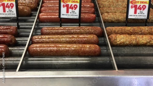 Sausages and hot dogs are spinning on a roller machine and displayed under bright retail lighting with deal pricing. 