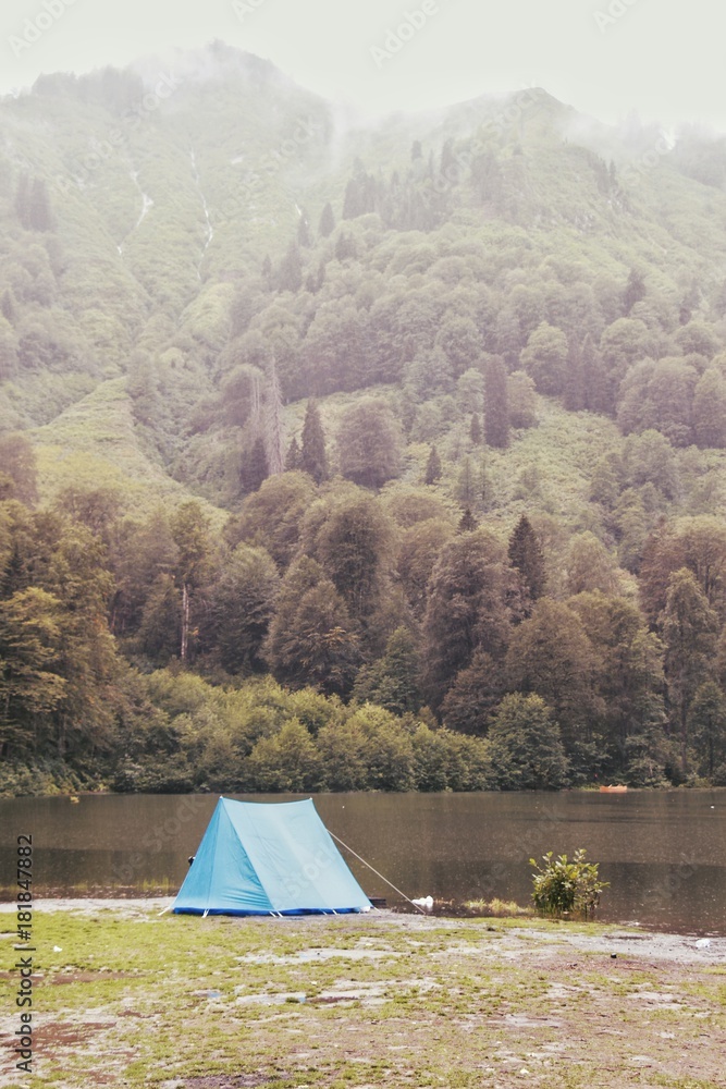 small cute blue tent on the lake shore background beautiful green forest in foggy weather