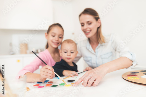Mother and children are engaged in drawing. They have fun in the kitchen. The girl is holding her younger brother in her arms.