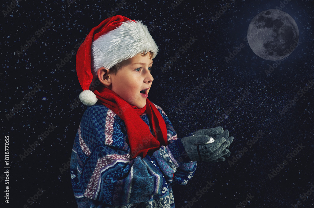 Happy boy in Santa hat plays with snowflakes on a dark background . Happy Christmas holidays Happy boy in Santa hat plays with snowflakes on a dark background . Happy Christmas holidays