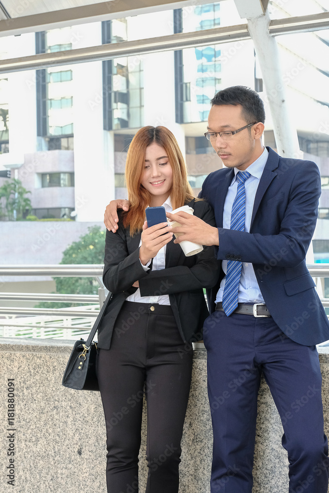 Businessman and businesswoman are holding a cup of coffee in break time during working and enjoy a smart phone at city area Businessman and businesswoman are holding a cup of coffee in break time during working and enjoy a smart phone at city area