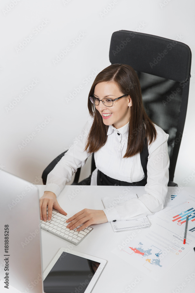 Beautiful smiling brown-hair business woman in suit and glasses sitting at the desk, working at computer with contemporary monitor with documents in light office, hands on keyboard on white background Beautiful smiling brown-hair business woman in suit and glasses sitting at the desk, working at computer with contemporary monitor with documents in light office, hands on keyboard on white background