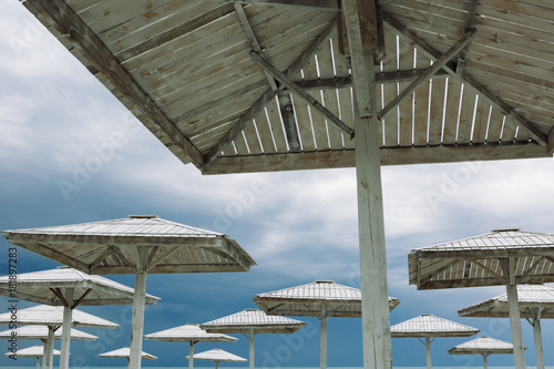 Fototapeta Naklejka Na Ścianę i Meble -  Wooden parasols and empty deckchairs on deserted beach on the off-season cloudy day