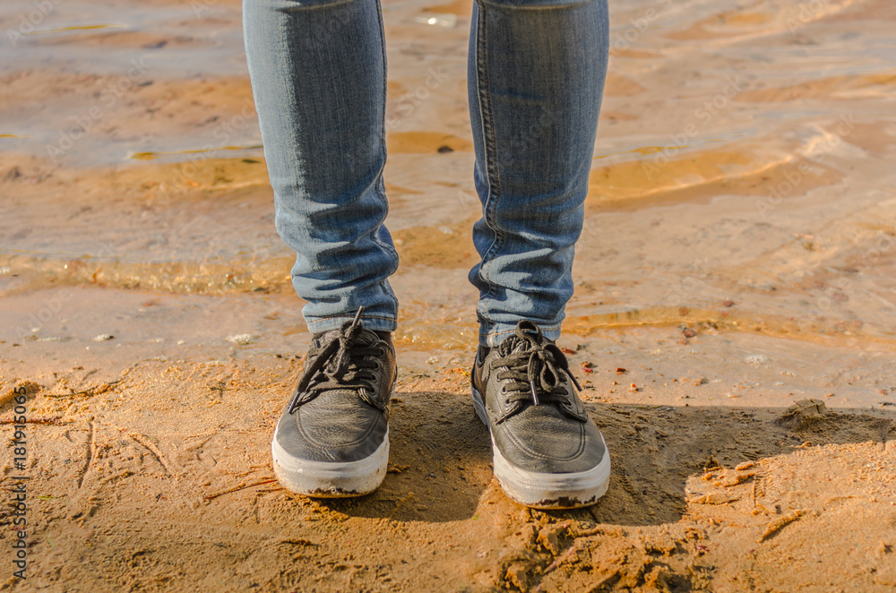 Female legs in jeans and sneakers on the beach - obrazy, fototapety, plakaty 