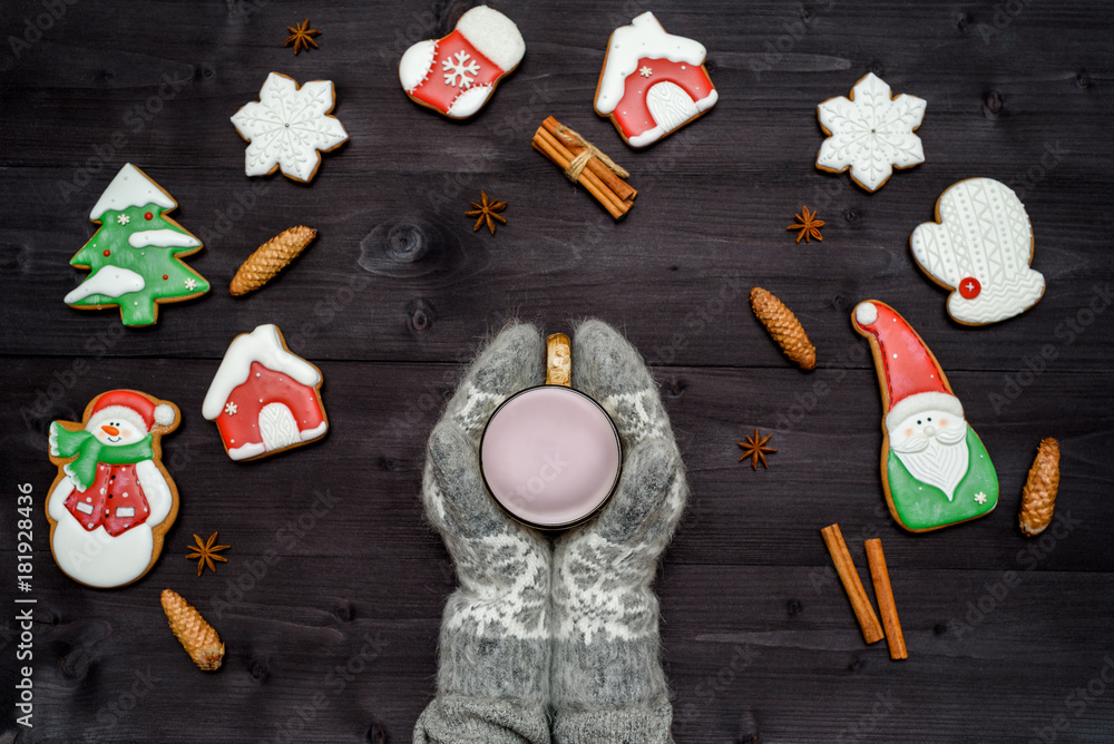 Woman hands in mittens holding cup of hot cocoa or chocolate on wooden table with homemade ginger cookies and cinnamon. Top view, flat lay. Christmas background. Cozy winter concept