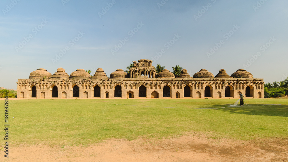 Ancient elephant stable building made in sandstone, in world heritage site of Hampi, Karnataka, India, with person watering the grass.