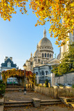 Rear view of the Basilica of the Sacred Heart of Paris in autumn at sunrise seen from a public park with orange leaves in the foreground against blue sky.
