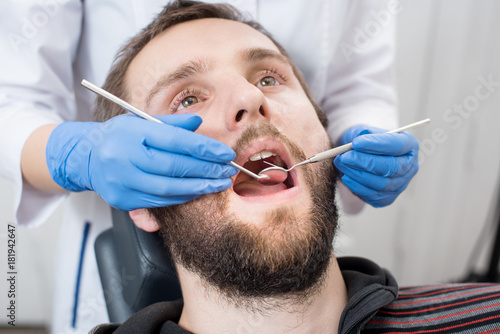 Close up of bearded man having dental check up in dental clinic. Dentist examining a patient s teeth with dental tools - mirror and probe. Dentistry.