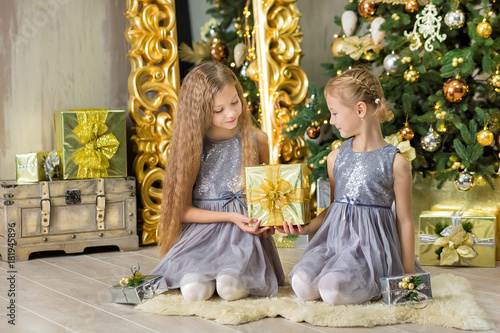 Merry Christmas and Happy Holidays Cute little child girls decorating the white green Christmas tree indoors with alot of presents wearing evening dresses with beautiful faces both