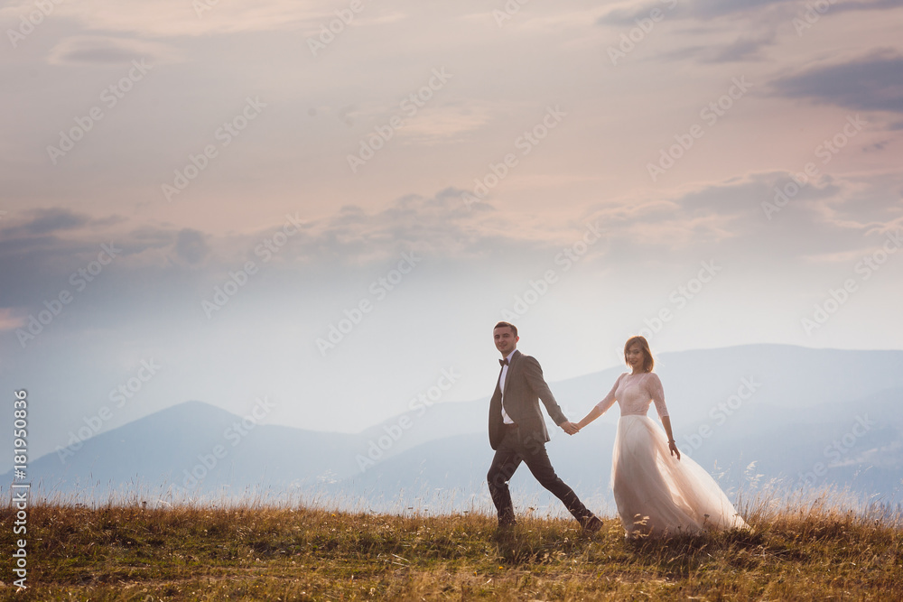 Fototapeta premium Bride and groom run on the hill before beautiful mountain landscape