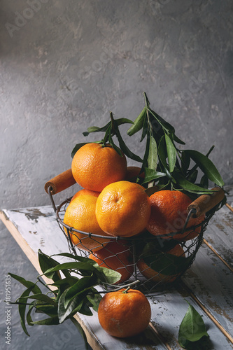 Ripe organic clementines or tangerines with leaves in basket standing on white wooden plank table with gray wall as background. Rustic style. Healthy eating. Toned image