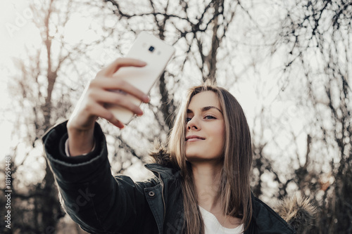 Pretty young woman taking selfie on mobile outdoor. Beautiful girl about twenty eyers old posing at camera in park. Model dressed in parka. Woman with blonde hair, full lips, wide eyebrows, blue eyes. photo