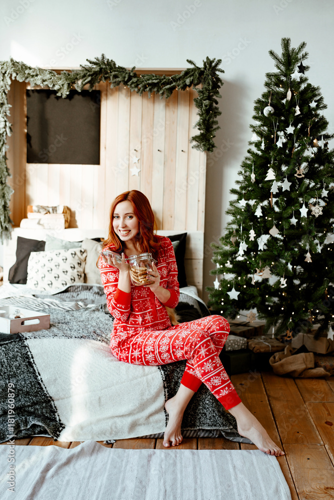 Fototapeta premium The sexy red-haired woman to a red pajamas has breakfast tea with home-made cookies in the bedroom which is beautifully decorated by Christmas against the background of the New Year's fir-tree.