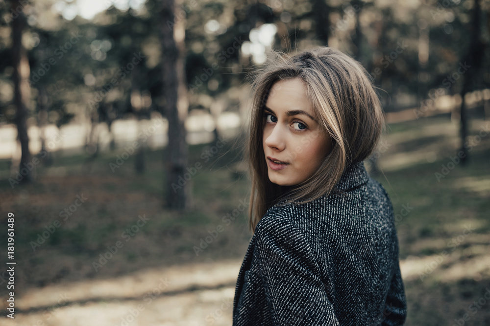 Gentle and pretty young woman with long blonde hair looking at camera over shoulder . Model posing at camera in coat in park outdoor Gentle and pretty young woman with long blonde hair looking at camera over shoulder . Model posing at camera in coat in park outdoor