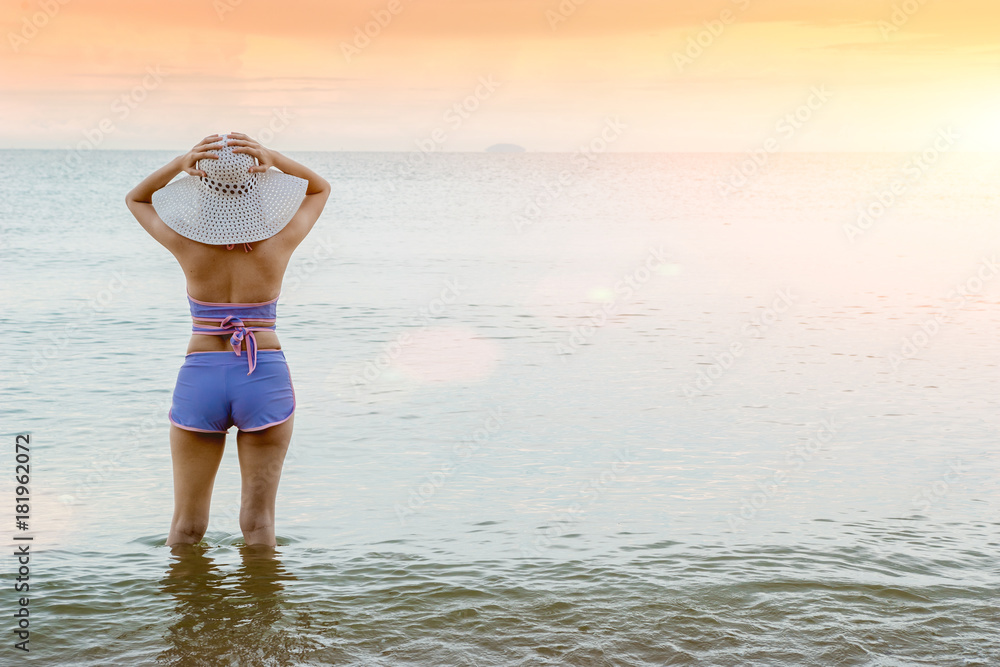 A young woman wearing a swimsuit and sunscreen white hat standing on a beautiful beach in front of the sunset on the day of the sea is beautiful. A young woman wearing a swimsuit and sunscreen white hat standing on a beautiful beach in front of the sunset on the day of the sea is beautiful.