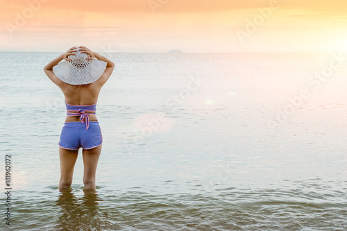 A young woman wearing a swimsuit and sunscreen white hat standing on a beautiful beach in front of the sunset on the day of the sea is beautiful.