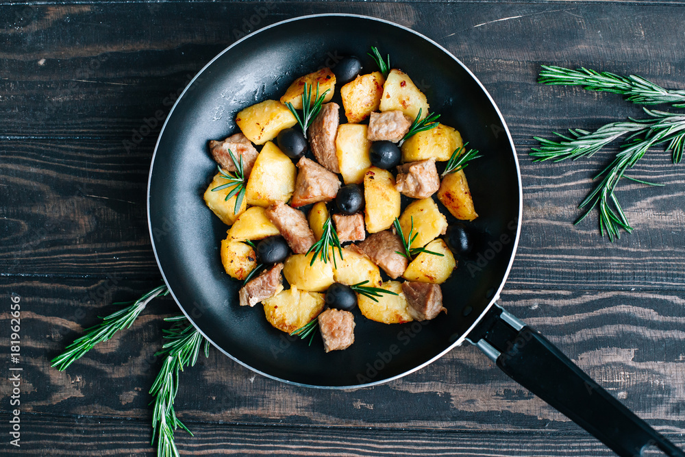 Fried potatoes with meat and olives decorated with rosemary in a frying pan on a wooden table top view Fried potatoes with meat and olives decorated with rosemary in a frying pan on a wooden table top view