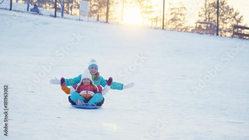 Family rides a sledding down on snow disk in sunny day against sunset