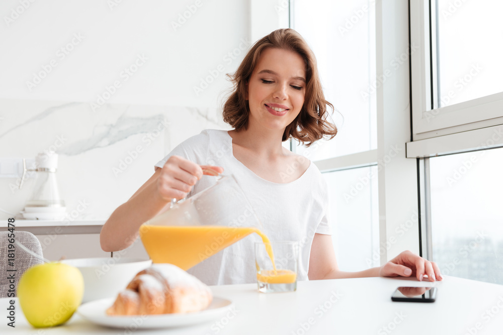 Cheerful brunette girl pouring juice into glass while sitting and having breakfast at the kitchen