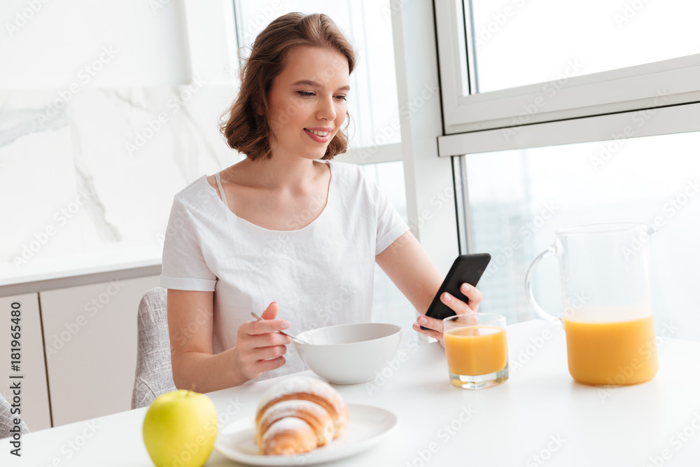 Young attractive woman in white tshirt using mobile phone while having breakfast at the kitchen Young attractive woman in white tshirt using mobile phone while having breakfast at the kitchen