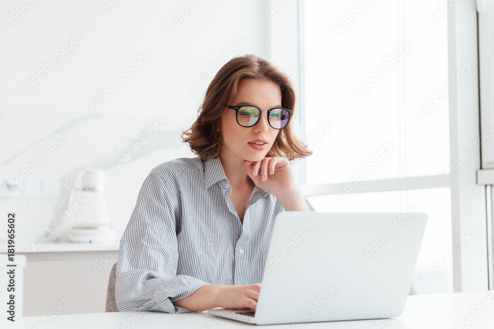 Charming businesswoman in glasses and striped shirt working with laptop computer while siting at home Charming businesswoman in glasses and striped shirt working with laptop computer while siting at home