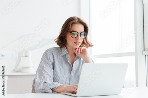 Charming businesswoman in glasses and striped shirt working with laptop computer while siting at home