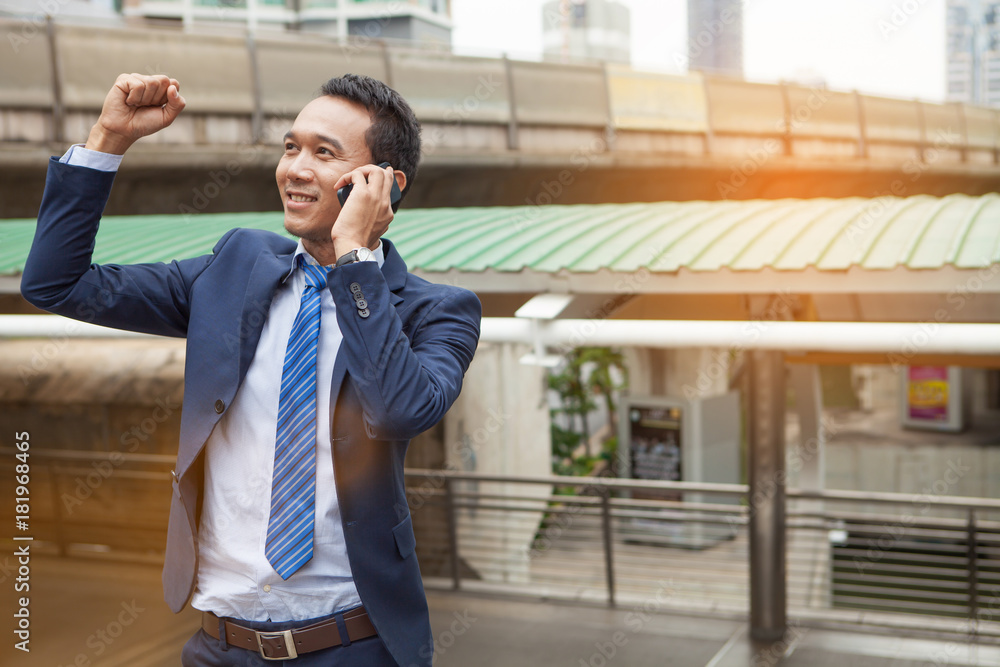 Businessman raising his fist in the air during callling the phone, with office building background - business success, achievement, and win concepts Businessman raising his fist in the air during callling the phone, with office building background - business success, achievement, and win concepts