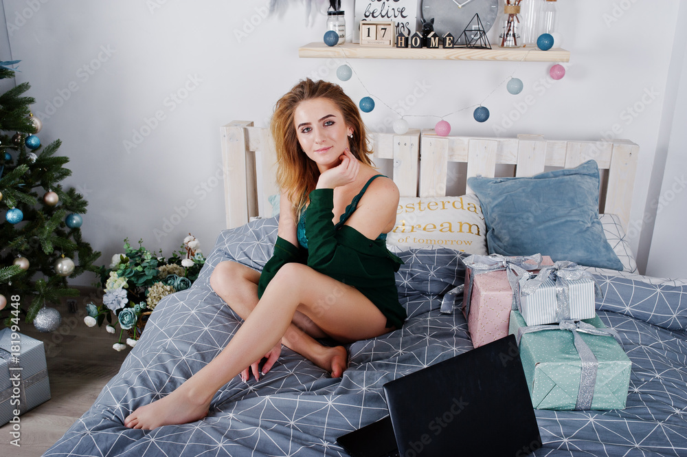 Sexy blonde model in green lingerie on bed with mobile phone and black laptop against new year tree.