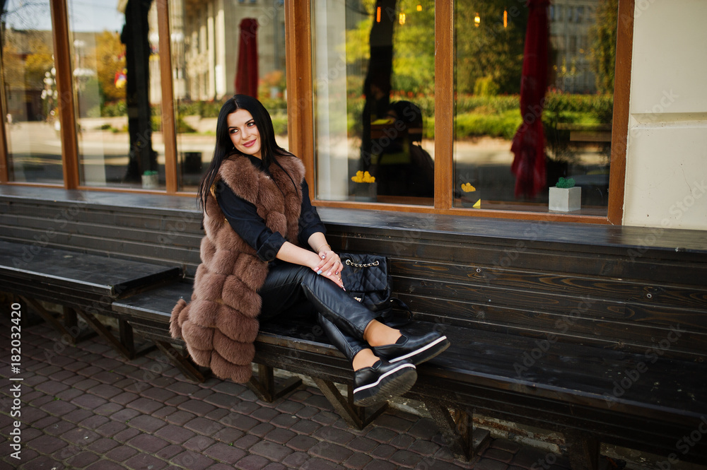 Fashion outdoor photo of gorgeous sensual woman with dark hair in elegant clothes and luxurious sleeveless fur coat and with backpack sitting on bench at autumn city.