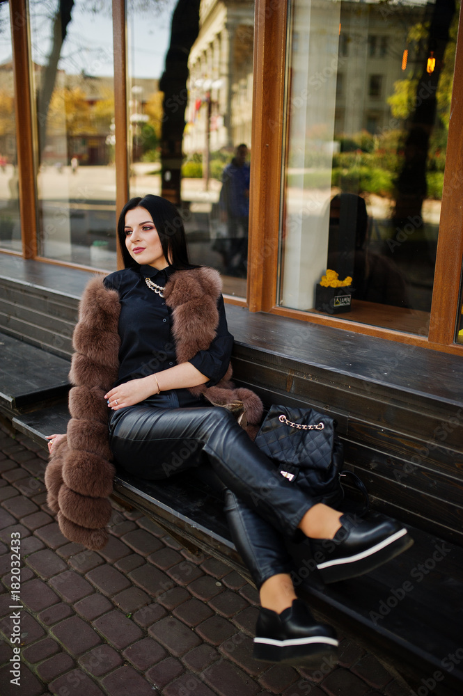 Fototapeta premium Fashion outdoor photo of gorgeous sensual woman with dark hair in elegant clothes and luxurious sleeveless fur coat and with backpack sitting on bench at autumn city.