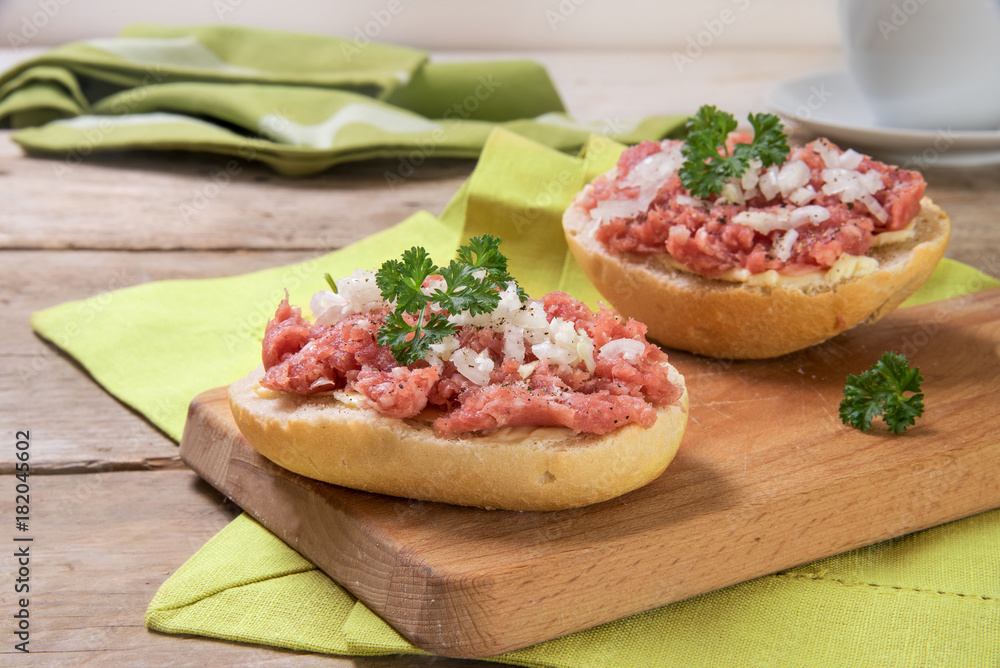 Buns with minced pork sausage, german mettwurst with onions and parsley garnish on a breakfast table with a wooden board and green napkins, close up