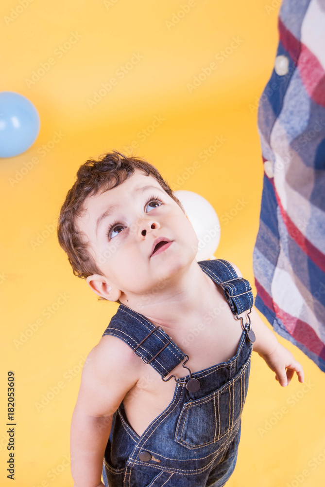 toddler in denim overalls on a yellow background with multi-colored balloons looks up at his father