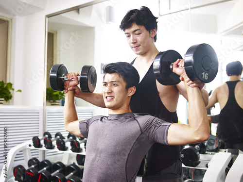 Canvastavla asian young adult working out in gym using dumbbells getting helped from trainer